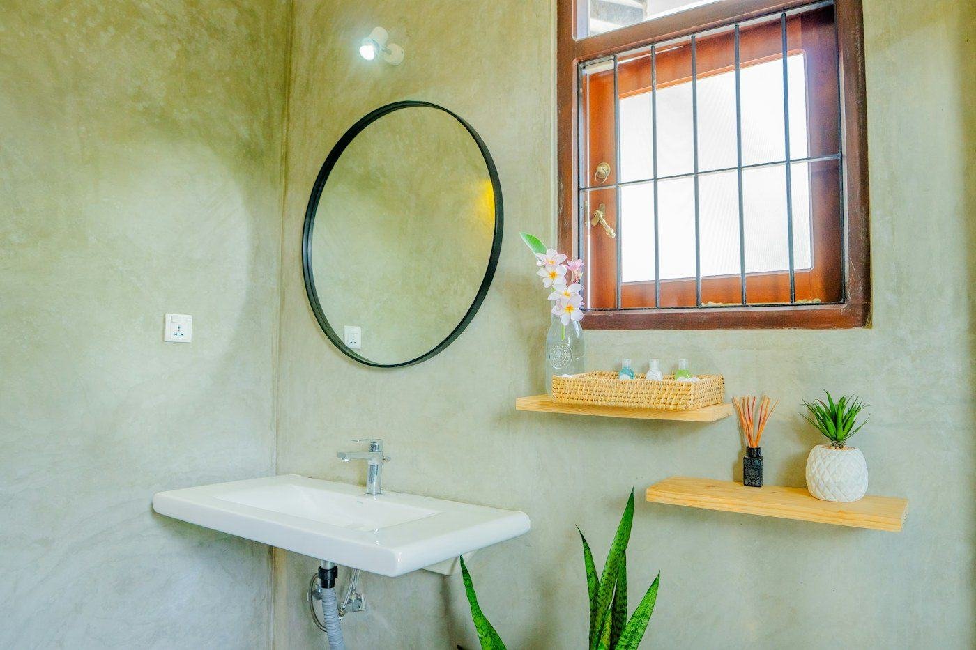A bathroom sink beneath a round mirror, with floating wooden shelves and potted plants.