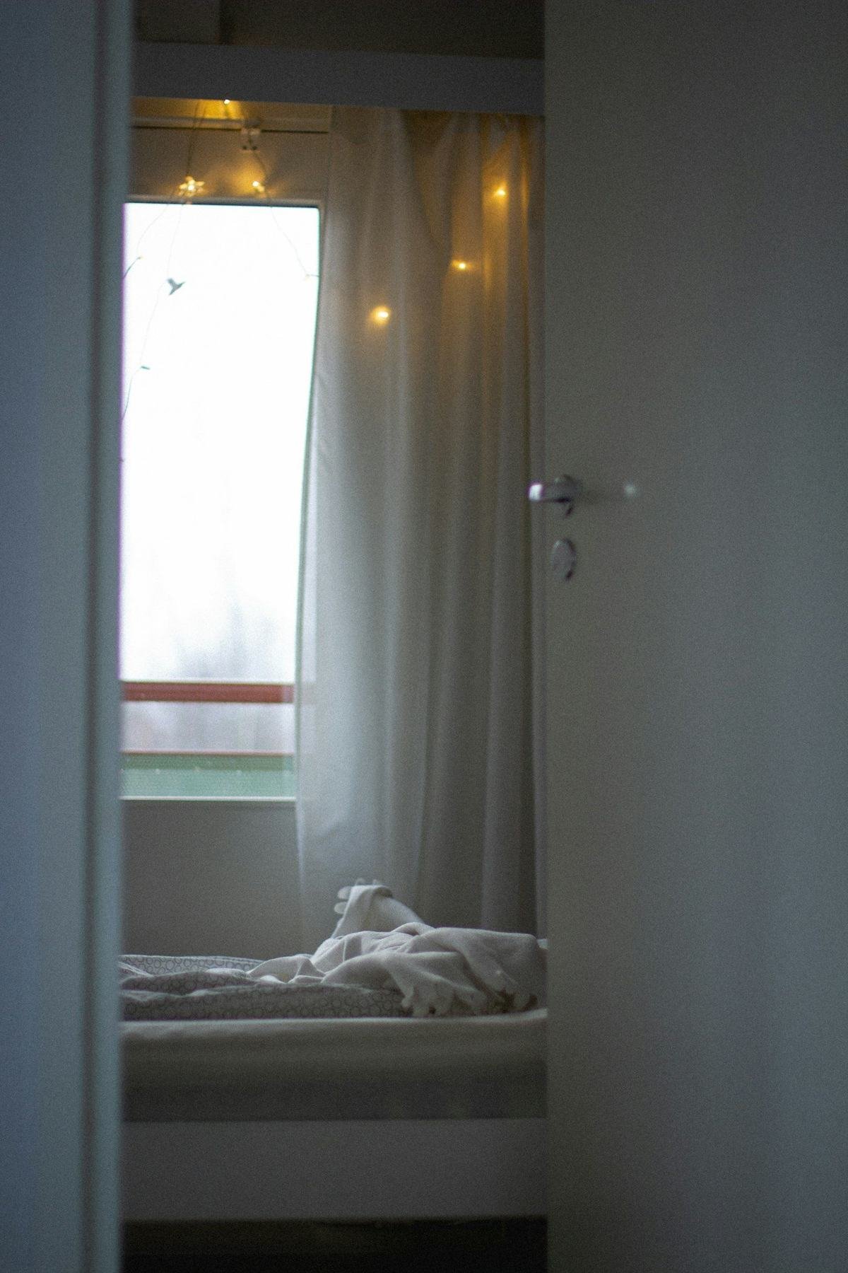 A narrow bedroom with a bed beside a window and pale curtains.