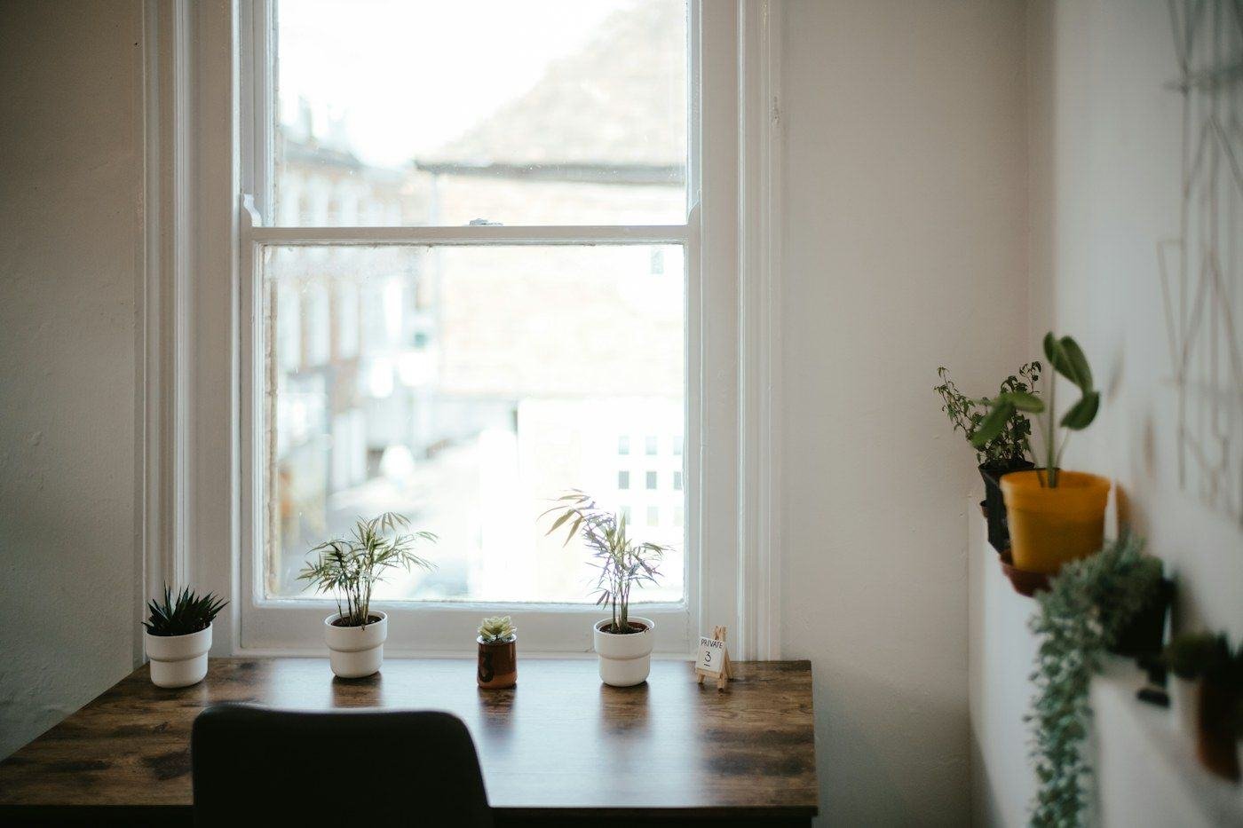 A wooden desk and dark chair beside a large window with small potted plants.