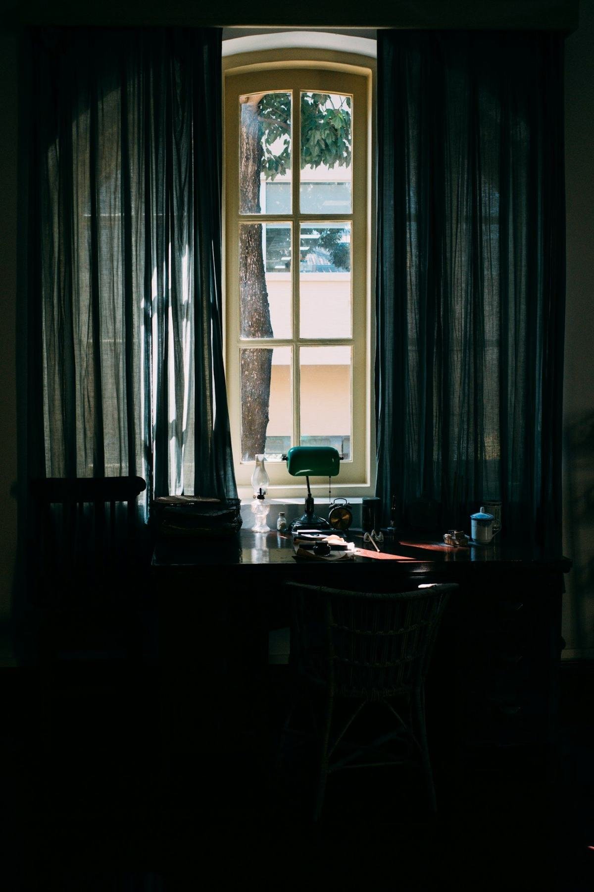 A small desk with books and a lamp beneath a tall window with dark curtains.