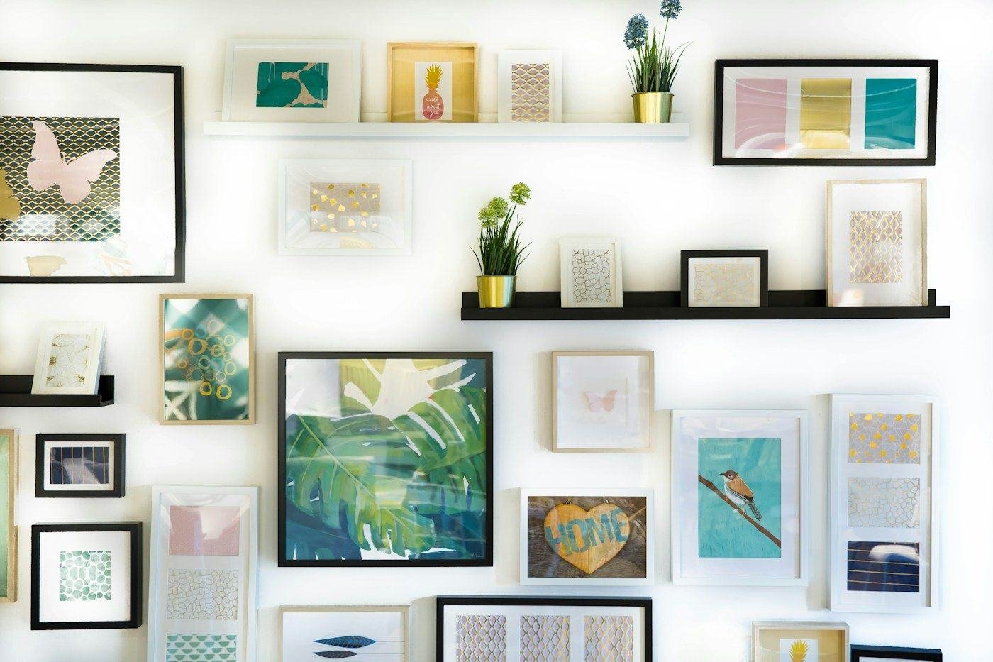 A collection of framed prints and small plants arranged on a white wall and picture ledges.