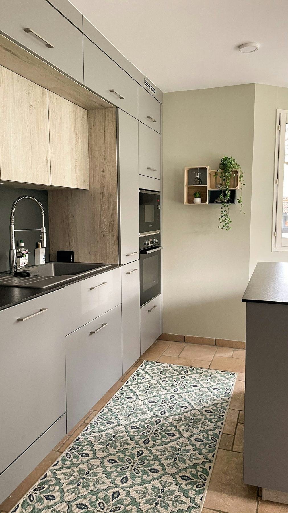 A narrow kitchen with pale wooden upper cabinets, white lower cabinets and a patterned runner.