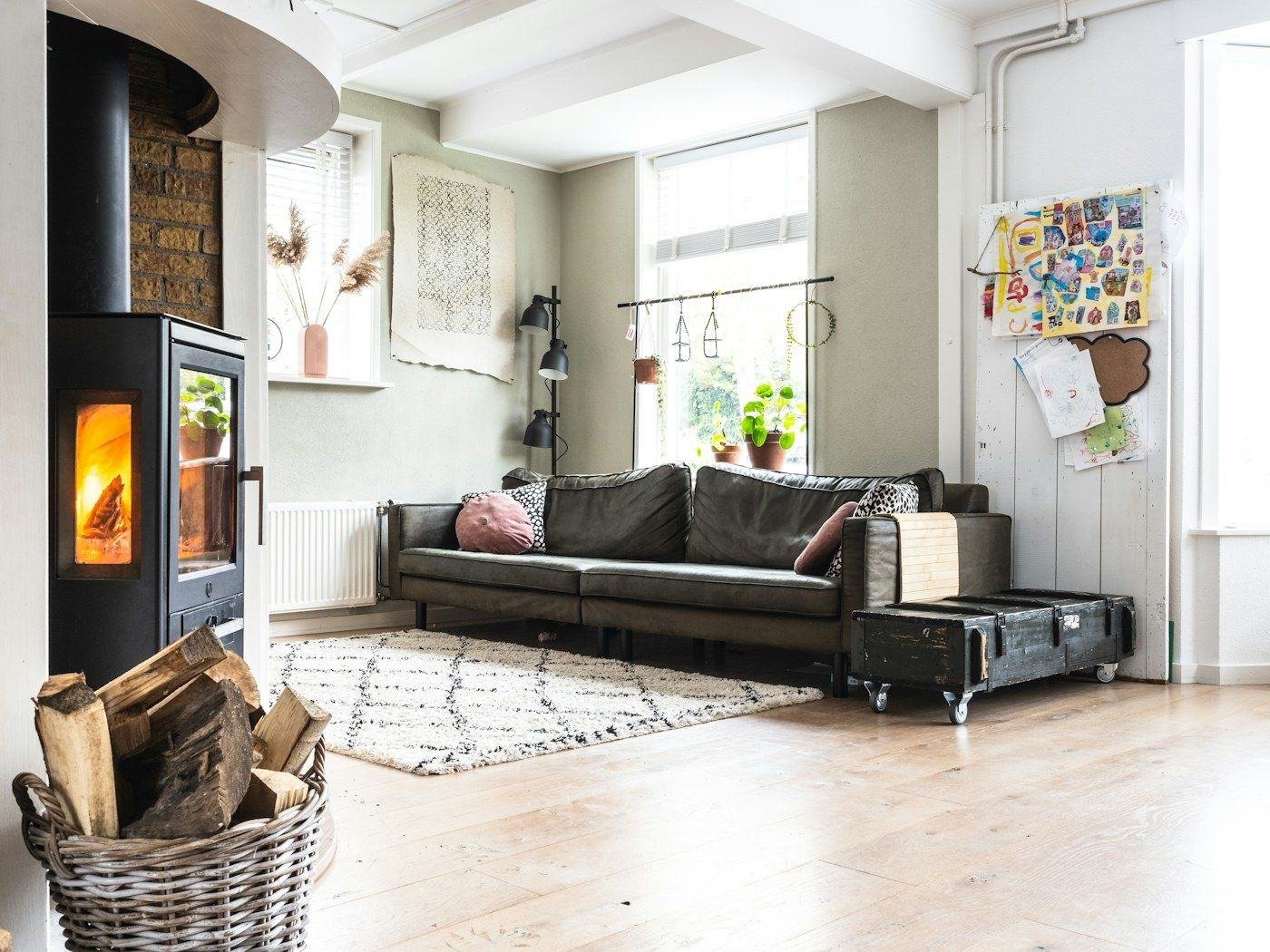 A dark sofa, pale rug and woven log basket in a bright room with a wood stove.